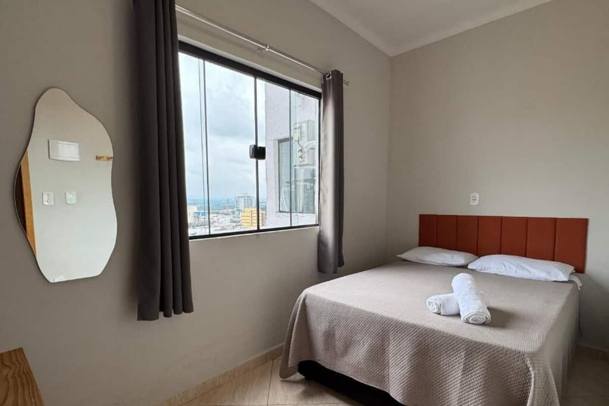 A hotel room with a bed, a window with curtains, a wall-mounted mirror, and a city view.