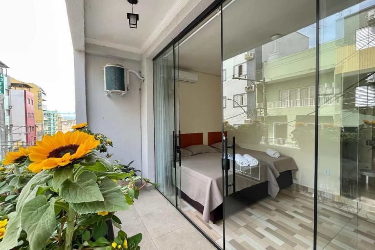 A hotel room with a bed, a glass door leading to a balcony, and a large potted plant with sunflowers.