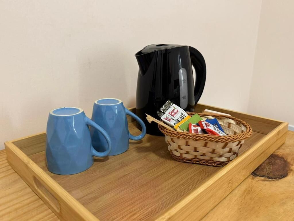 A tray with two blue mugs, a black coffee pot, and a basket of coffee packets on a wooden surface.