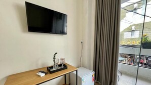 A room with a flat-screen TV mounted on the wall, a small white refrigerator, a wooden console table with a remote control, a glass vase, and a grey cushion.