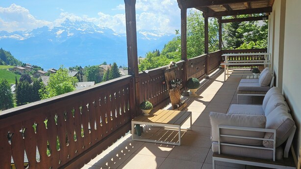 A covered balcony with a view of mountains, outdoor furniture, and a wooden railing. - Spacious central 5-room apartment (Leysin)