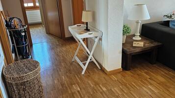 Een kamer met een houten vloer, een witte inklapbare tafel met een lamp, een rieten mand en een donkergrijze bank.
