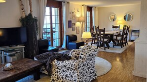 A living room with a sofa, a TV, a dining table with chairs, and a fireplace. - Spacious central 5-room apartment (Leysin)