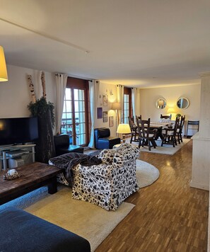 Een woonkamer met een bank, een televisie, een eettafel met stoelen en een open haard.