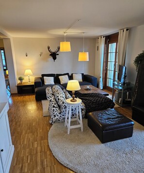 Een woonkamer met een zithoek, een salontafel, een lamp, een decoratief hertenhoofd en een groot raam.