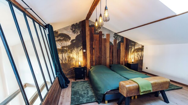 Un dormitorio con techo inclinado, cama con colcha verde, banco de madera y mesita de noche con lámpara.