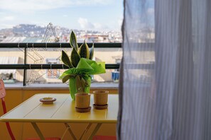 A table with a potted plant and two cups, a red chair, and a view of a cityscape through a window with a curtain. - Seven Nap - Central Rooms (Naples)