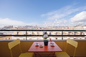 Uma varanda com uma mesa vermelha, duas cadeiras amarelas, um vaso com flores brancas e vista para um horizonte urbano com edifícios e uma ponte.