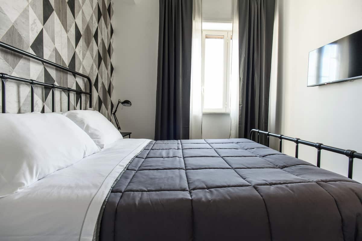Una cama con colcha gris, una pared con diseño geométrico, una ventana con cortinas y un televisor montado en la pared.