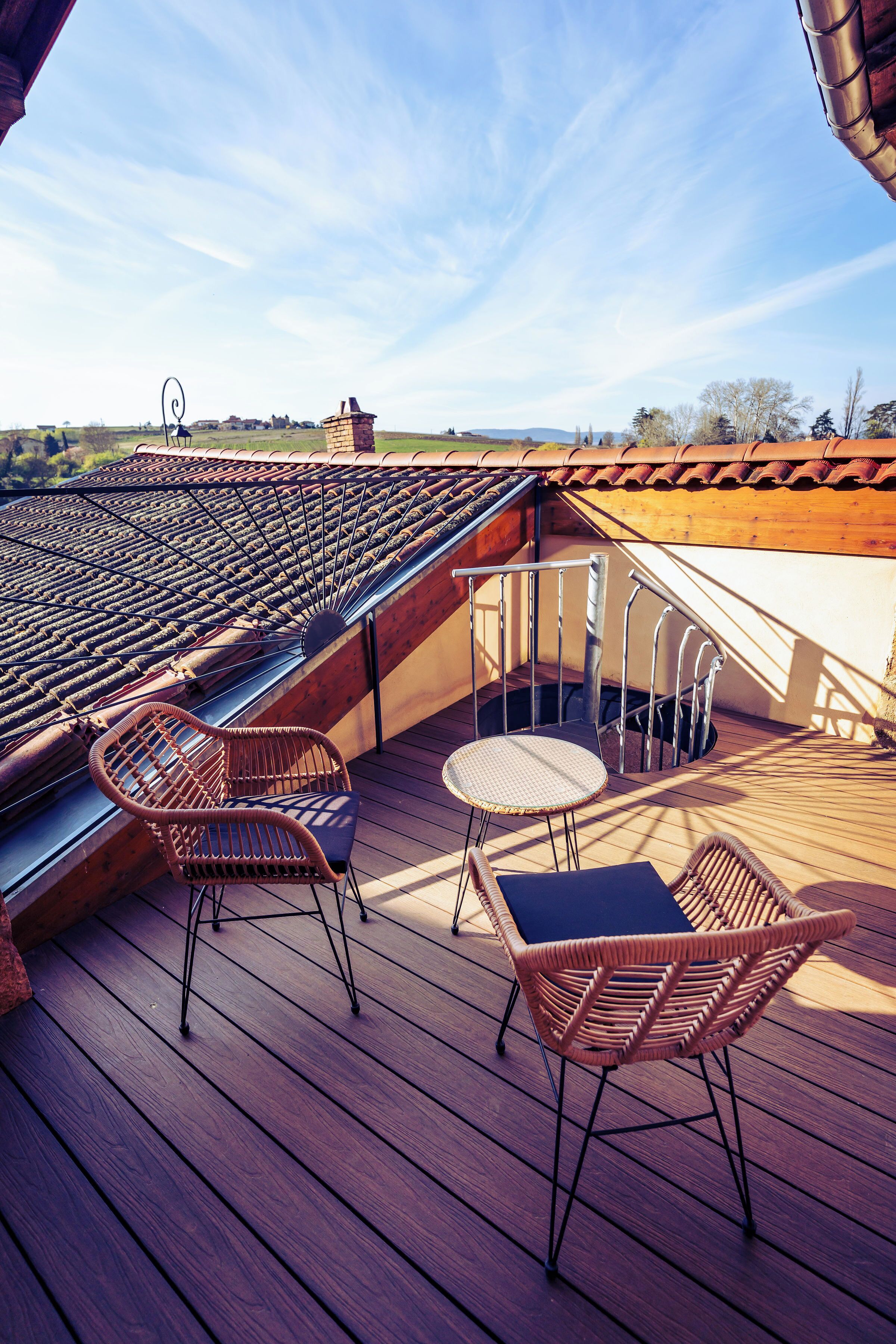 Une terrasse en bois avec des chaises et une table en osier, un toit en tuiles et une vue sur la campagne.