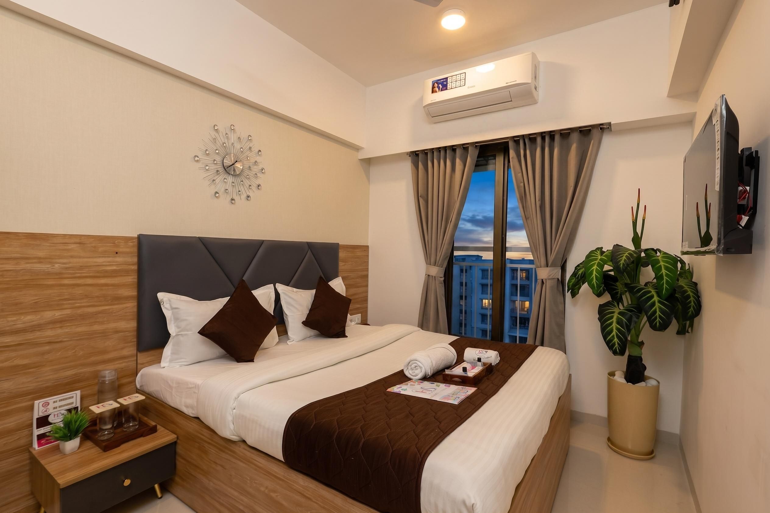 A hotel room with a bed, bedside table, air conditioning unit, TV, window with curtains, and a potted plant.