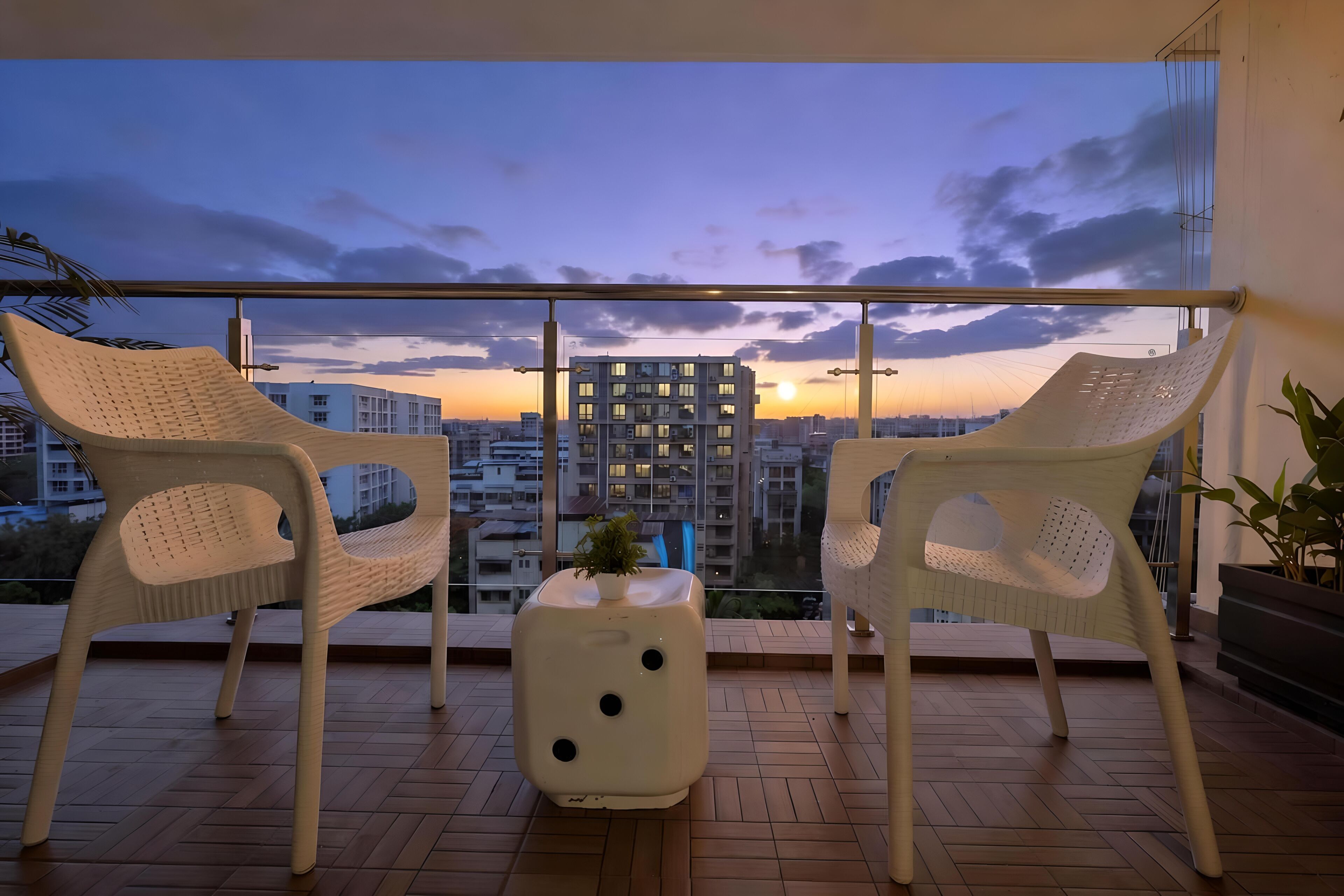 A balcony with two white chairs and a small table with a plant, overlooking a cityscape at sunset.