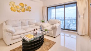 A modern living room with a sofa set, a glass coffee table, a decorative wall piece, a potted plant, and a large window with curtains. - Santacruz Homesuites - Gagal Home (Mumbai)