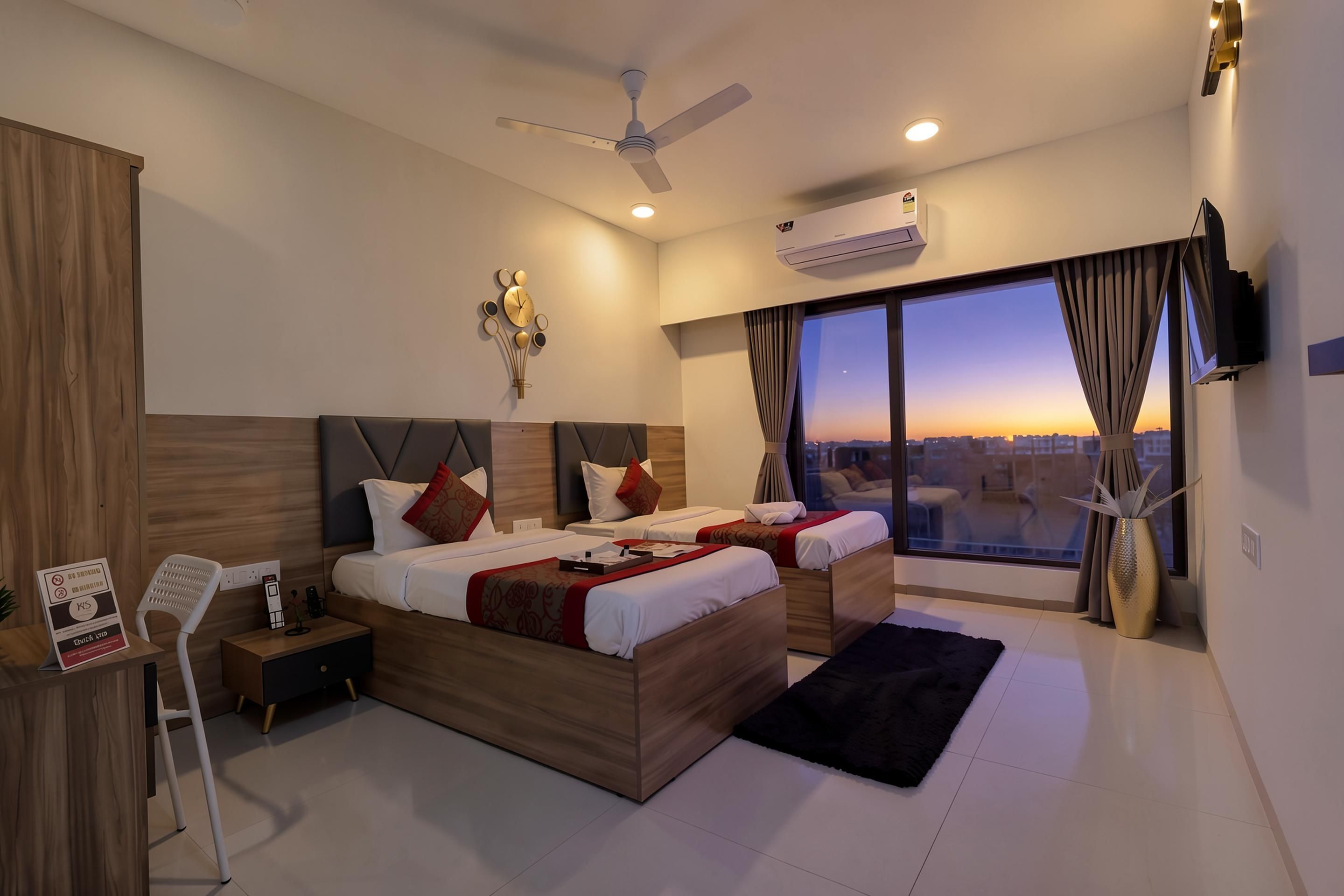 A modern hotel room with two beds, a desk, a chair, a ceiling fan, an air conditioning unit, and a large window with a city view.