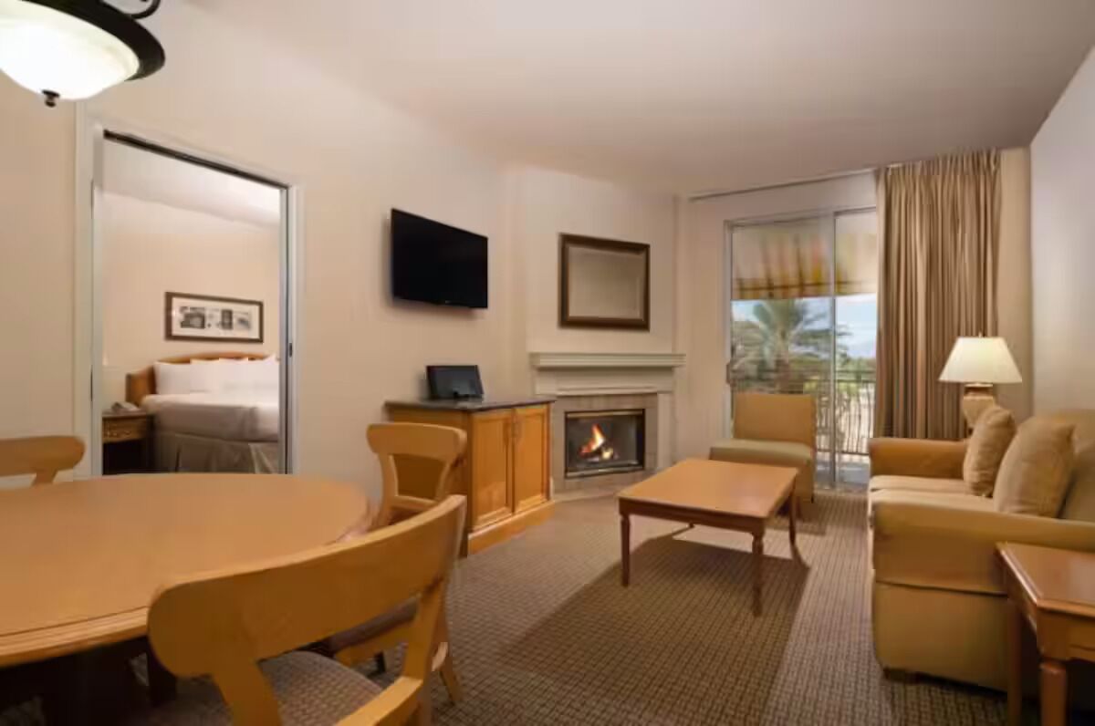 Habitación de hotel con mesa de comedor, sillas, sofá, televisor, chimenea y una ventana grande con cortinas.