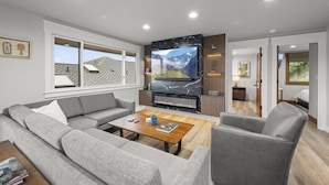 A modern living room with a large flat-screen TV, grey sofas, a wooden coffee table, and a fireplace.