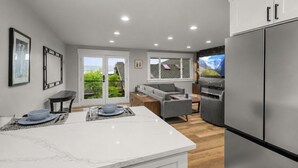 A modern kitchen with a large refrigerator, a dining area with a table and chairs, and a living area with a sofa and TV.