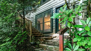 Una escalera de piedra que lleva a una puerta de madera, rodeada de abundante vegetación y un edificio con ventanas grandes.