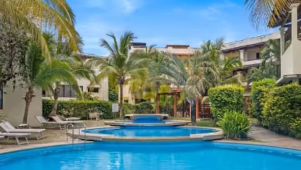 Una piscina con sillones, palmeras y un edificio con balcones al fondo.