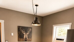 A kitchen with a wooden table, two stools, a hanging light fixture, a wall-mounted air conditioner, and a window with a view. - This house is a 2 bedroom(s), 2 bathrooms, located in Kalkaska, MI. (Kalkaska)