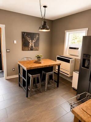 A kitchen with a wooden table, two stools, a hanging light fixture, a wall-mounted air conditioner, and a window with a view.