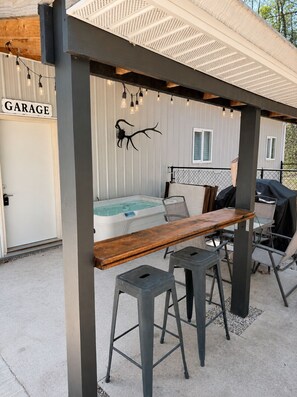 An outdoor patio with a wooden table, two stools, a hot tub, and a grill.