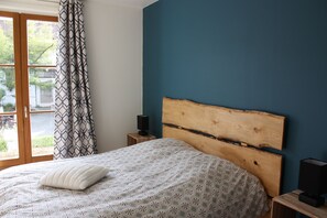 Ein Schlafzimmer mit einem Bett, einem Holz-Kopfteil, einem Nachttisch mit Lautsprecher und Blick ins Freie durch ein Fenster.