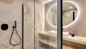 Ein modernes Badezimmer mit einer gläsernen Duschkabine, einem runden Spiegel mit integrierter Beleuchtung und einer Wand mit Marmorfliesen.