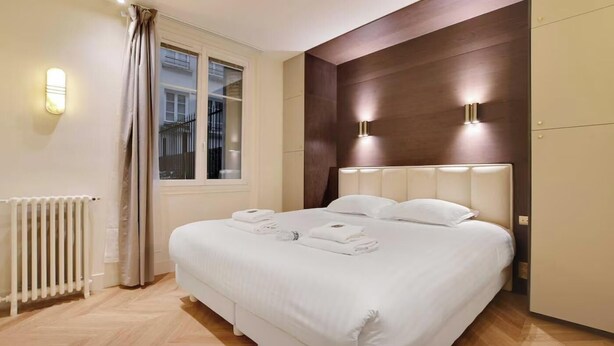 A hotel room with a large bed, a radiator, a window with curtains, and wall-mounted lighting. - AN AUTHENTIC PARISIAN LIVING AT BLVD SAINT GERMAIN (Paris)