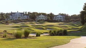Ein Golfplatz mit sanften grünen Hügeln, Sandbunkern und einem Weg, der zu einer Reihe großer, weißer, im Kolonialstil erbaute Gebäude führt.