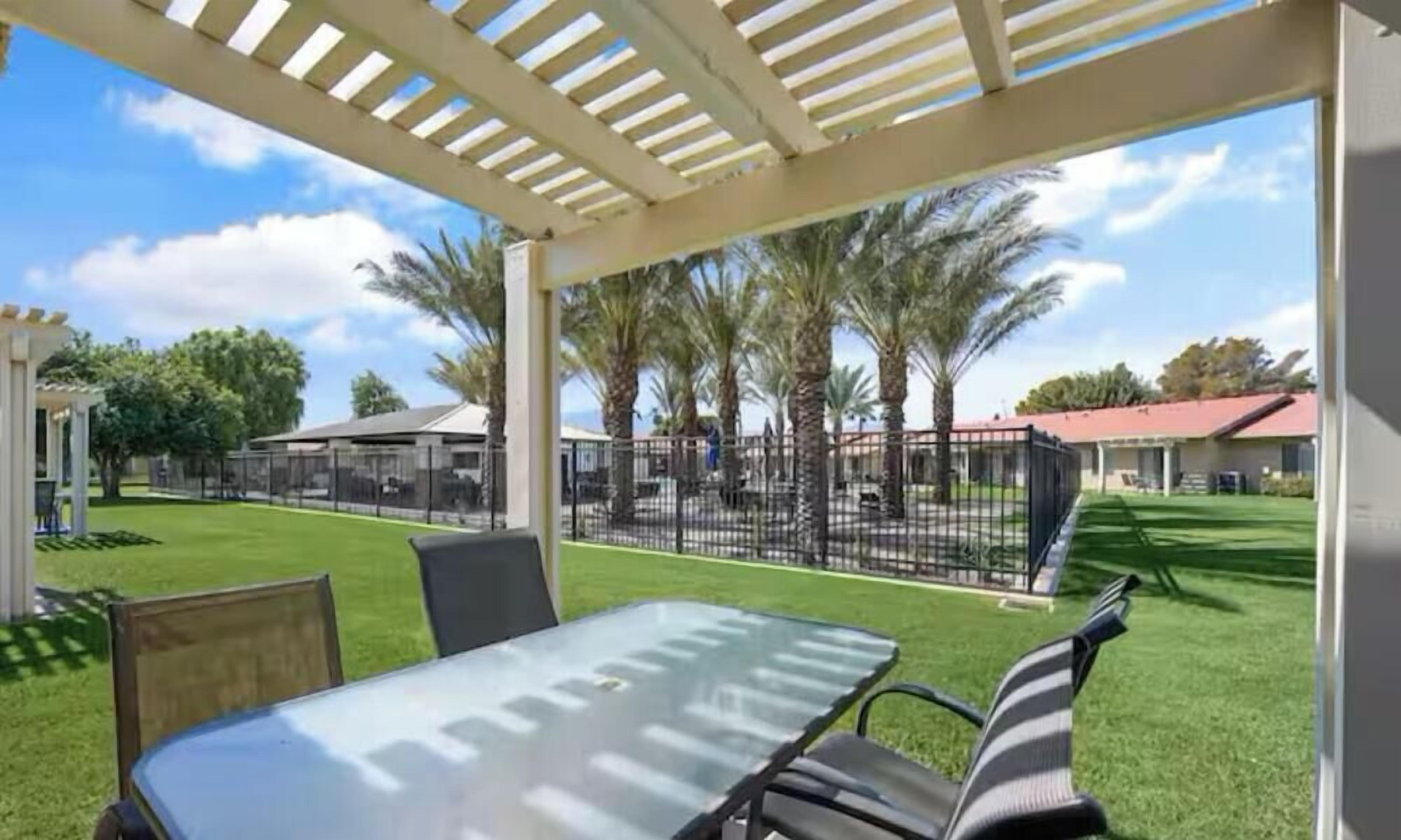 A covered outdoor area with a table and chairs, surrounded by a fence and palm trees.