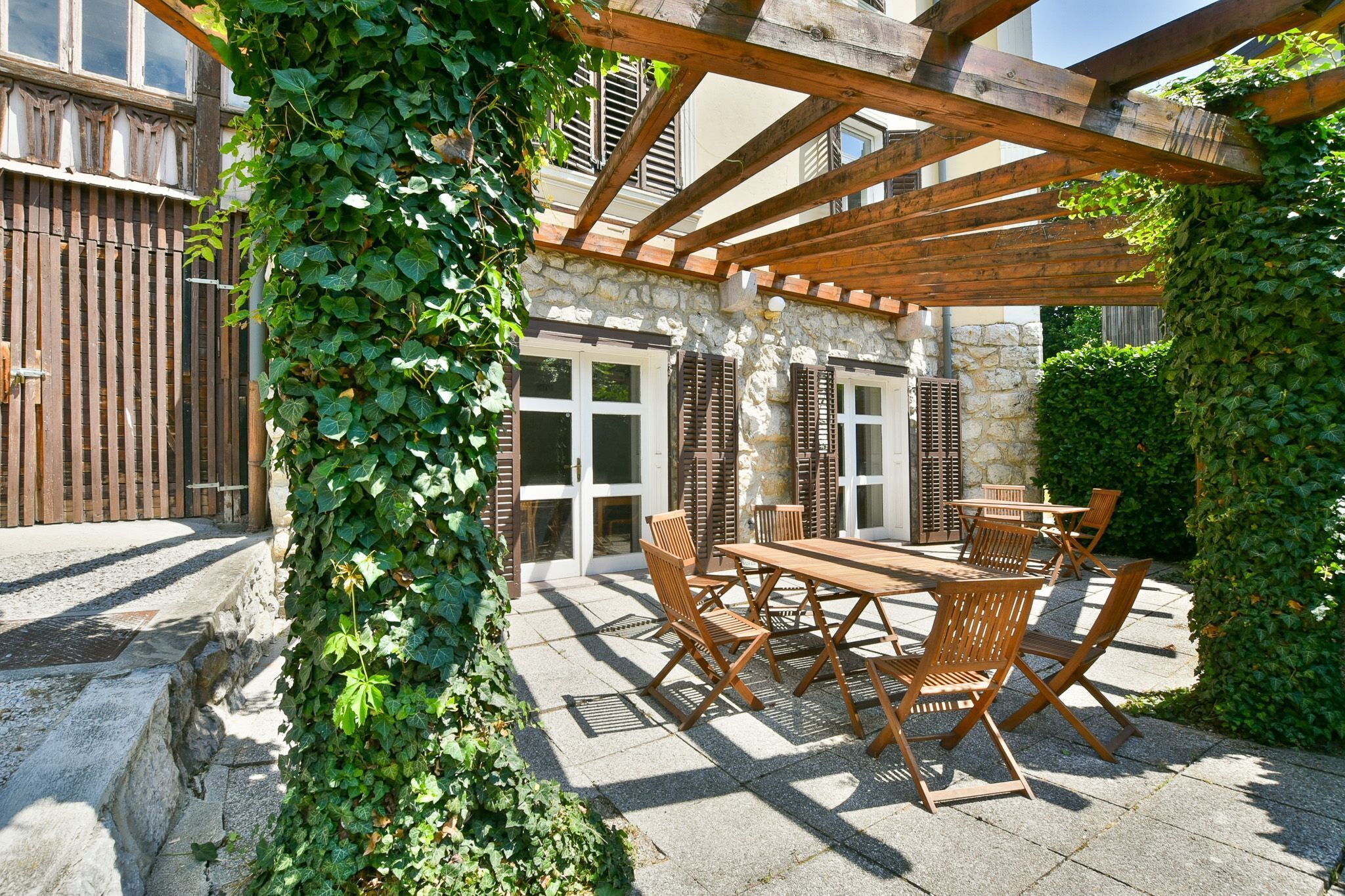 An outdoor patio with a wooden pergola, stone walls, and a table with chairs.