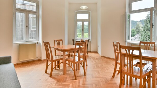 Un espace repas comprenant une grande table et des chaises, de grandes fenêtres donnant sur la verdure et un luminaire au plafond.