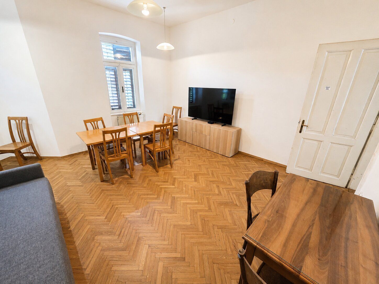 A room with a herringbone wooden floor, a dining table with chairs, a television on a cabinet, and a door.