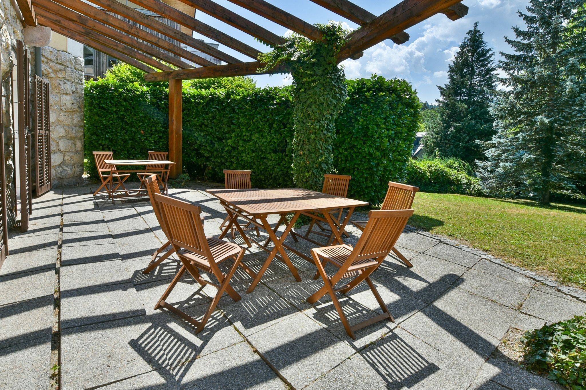 An outdoor patio with a pergola, wooden tables and chairs, stone flooring, and a hedge in the background.