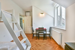 Ein Zimmer mit Holzboden, einem Esstisch mit Stühlen, einer weißen Treppe und einem Fenster mit Jalousien.