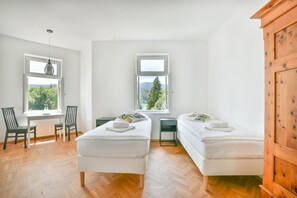 Ein Zimmer mit zwei Betten, einem Schreibtisch und zwei Stühlen. Fenster bieten Blick auf Bäume und Berge.