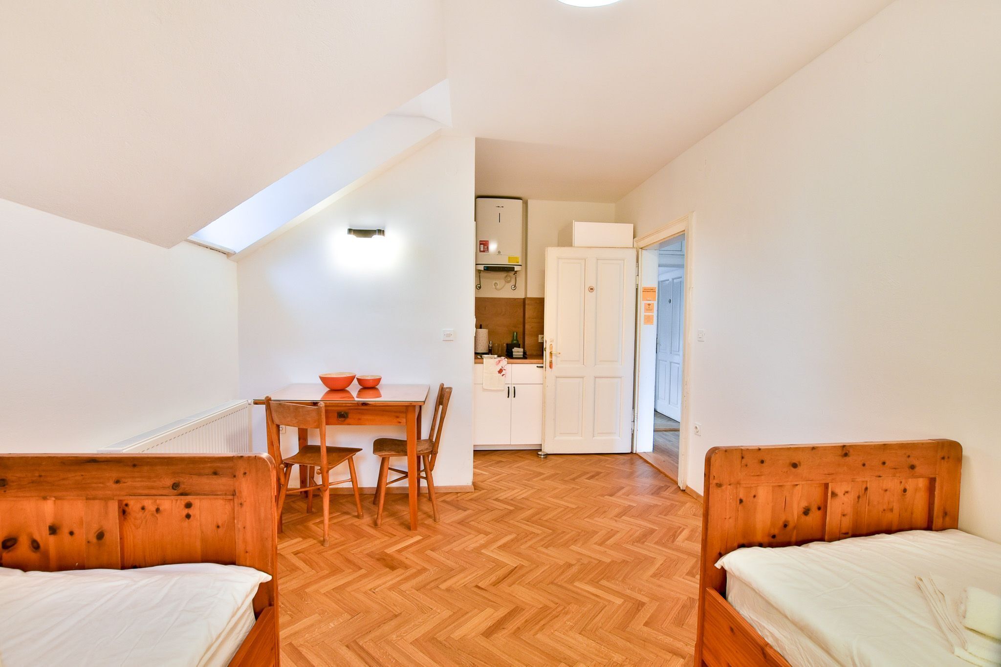 A small, compact room with a wooden floor, a bed, a table with chairs, and a kitchenette in the background.