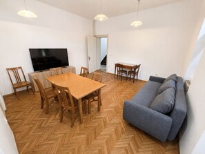 Ein Wohnzimmer mit Holzdielen-Fischgrätmuster, einer grauen Couch, einem Holzesstisch mit Stühlen, einem Flachbildfernseher und Pendelleuchten.
