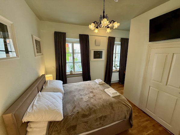 A bedroom with a bed, a chandelier, a window with curtains, a door, and a radiator.