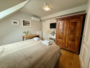 A bedroom with a bed, a vanity with a mirror, a wooden wardrobe, and a desk with a vase of flowers.