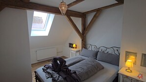 A bedroom with a sloped ceiling, a skylight, a bed with striped bedding, a bedside table with a lamp, and a radiator. - Historic horse stable in the center of Weißenburg (Weißenburg in Bayern)