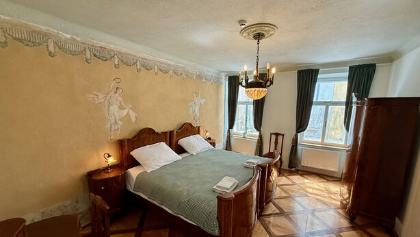 A room with a bed, a chandelier, a window with curtains, and a radiator. - Mini-Teater Suites - Apartment 1  - sleeps 3 guests  in 1 bedroom (Ljubljana)