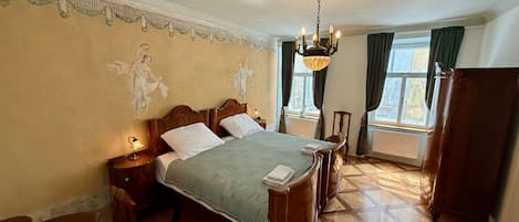 Une pièce avec un lit, un lustre, une fenêtre avec des rideaux et un radiateur.