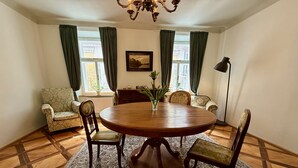 A dining room with a round wooden table, four chairs, a chandelier, a window with curtains, a radiator, and a framed picture on the wall. - Mini-Teater Suites - Apartment 1  - sleeps 3 guests  in 1 bedroom (Ljubljana)
