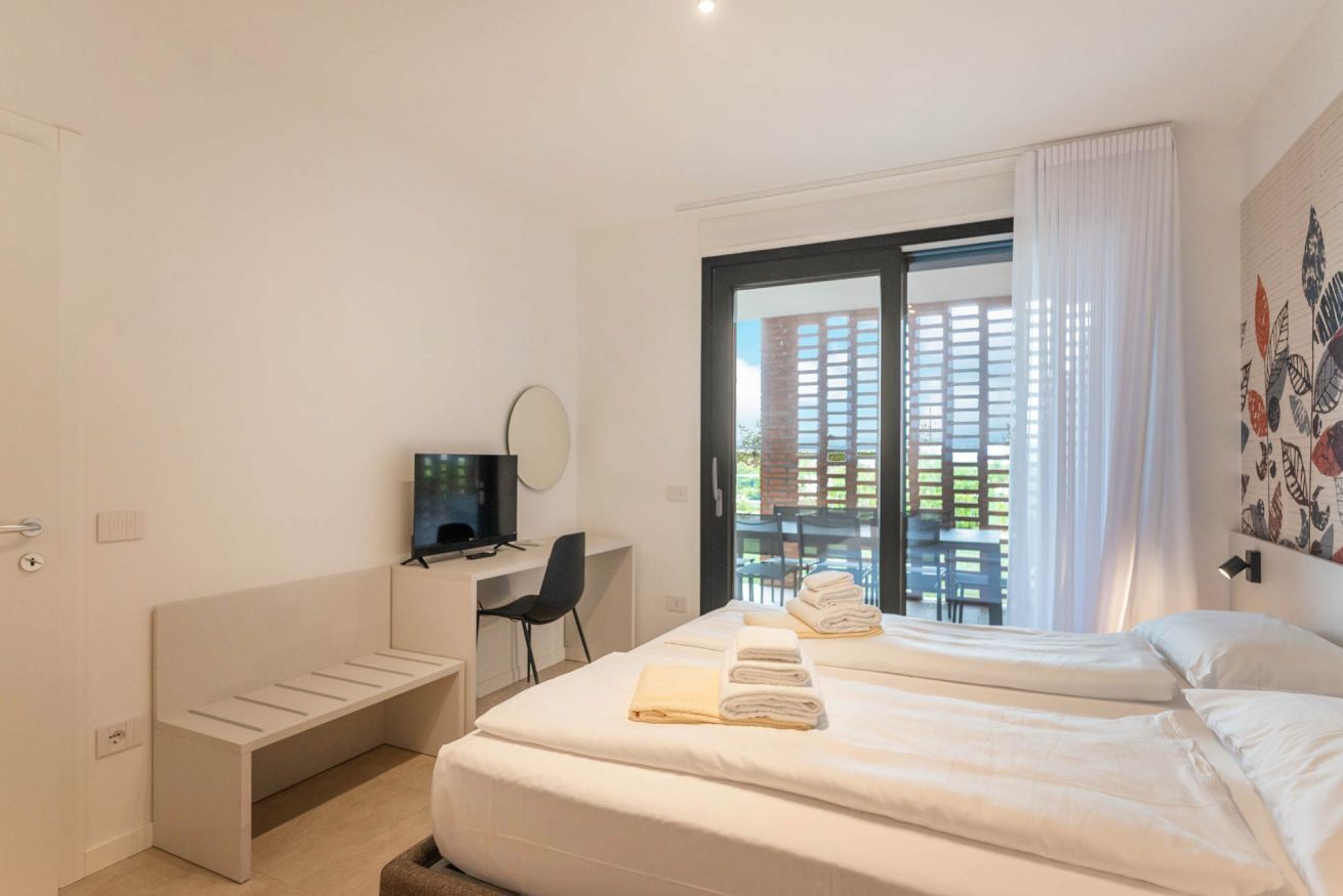 A hotel room with a bed, a desk, a television, and a view of a building through the window.
