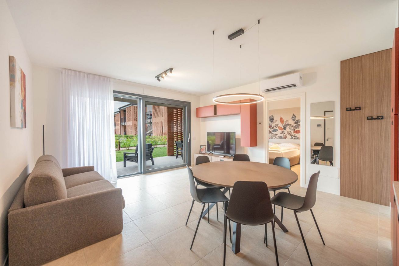 A modern living room with a sofa, a dining table with chairs, a TV, and a sliding glass door leading to an outdoor area.