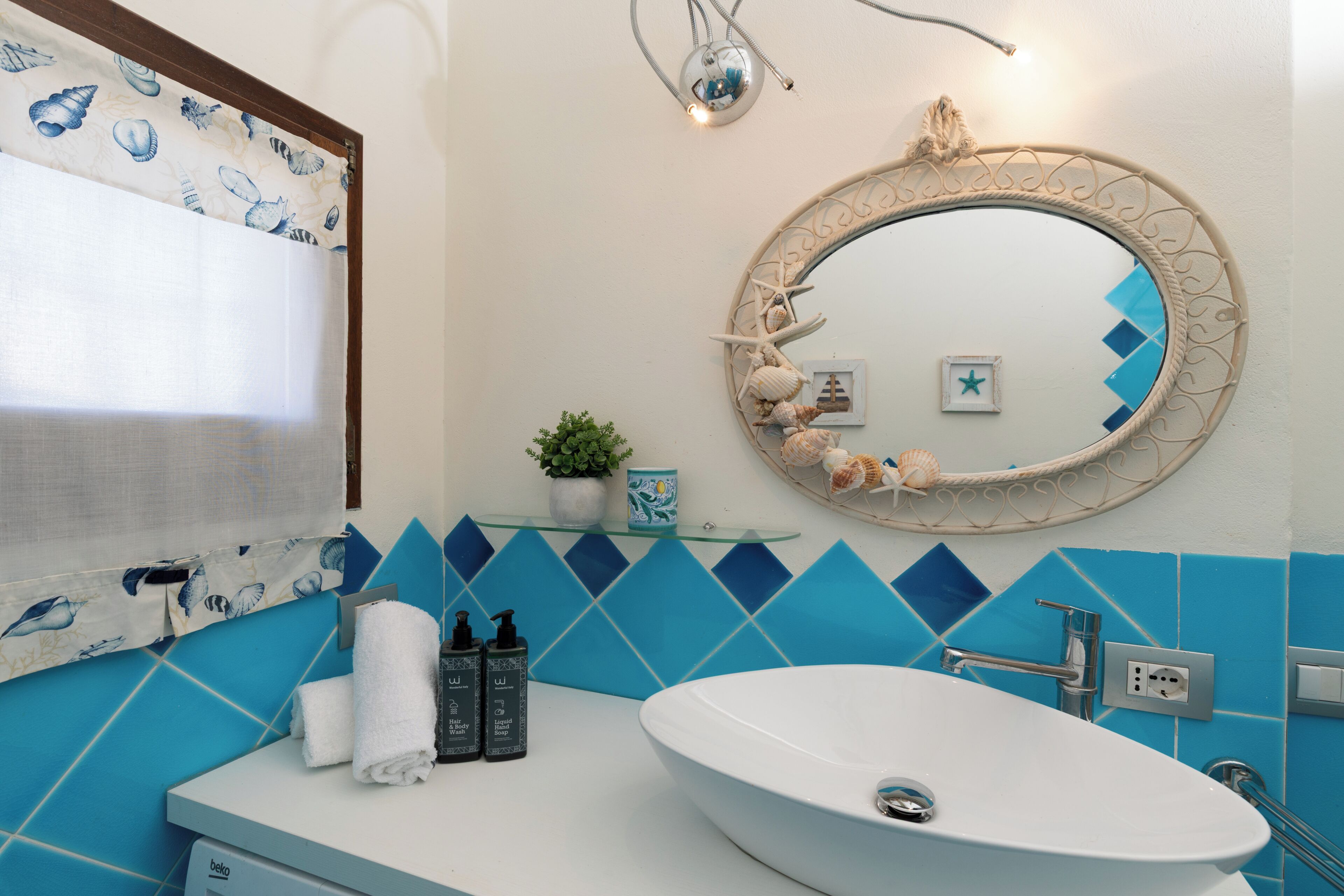 Een badkamer met een witte wastafel, blauwe tegels, een ronde spiegel met decoratieve details en een raam met gordijn.