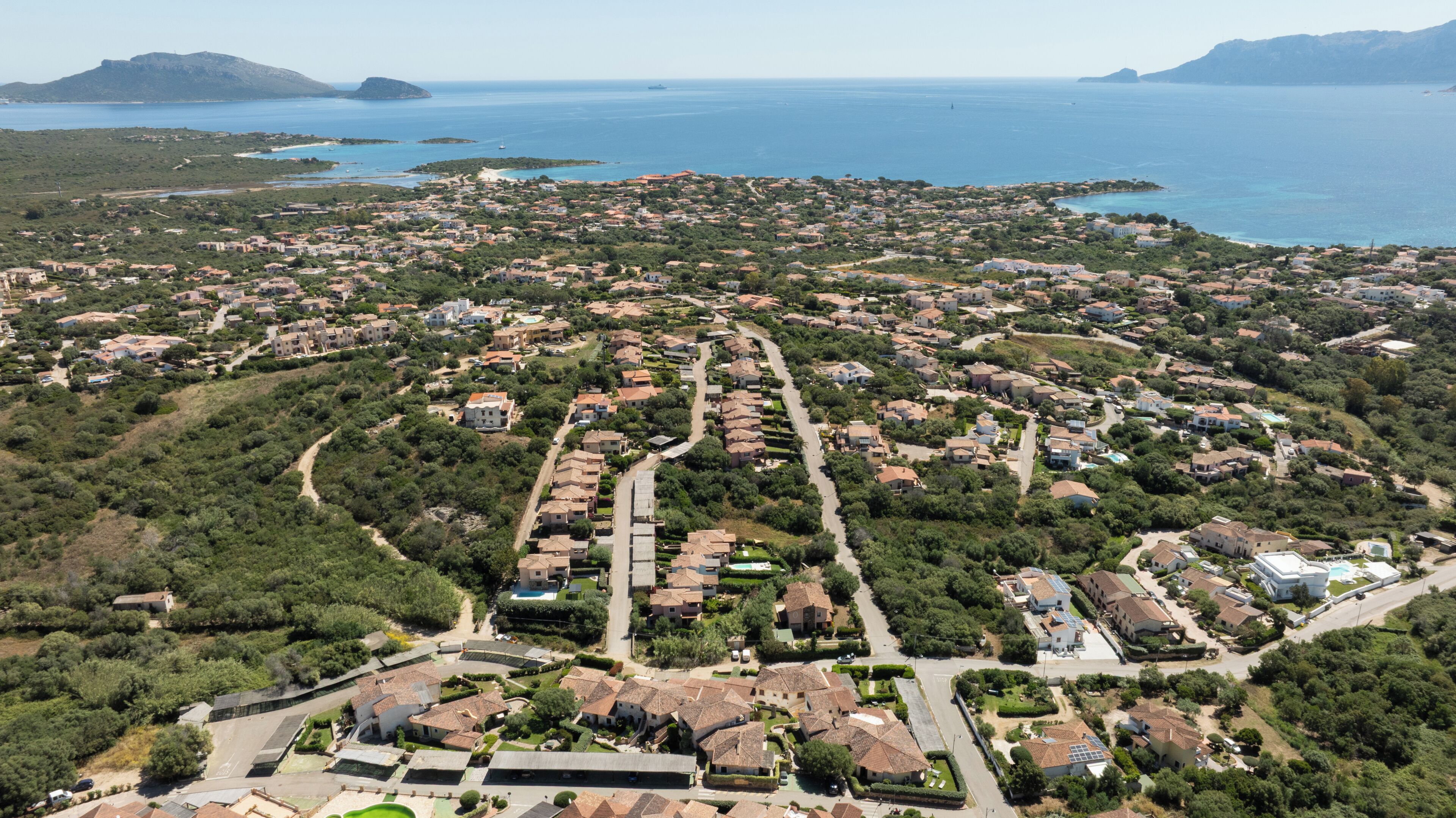 An aerial view of a coastal residential area with houses, roads, and a clear view of the sea and distant islands.