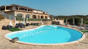 Una piscina con solárium de piedra, mobiliario exterior y un edificio con arcos y fachada de piedra.